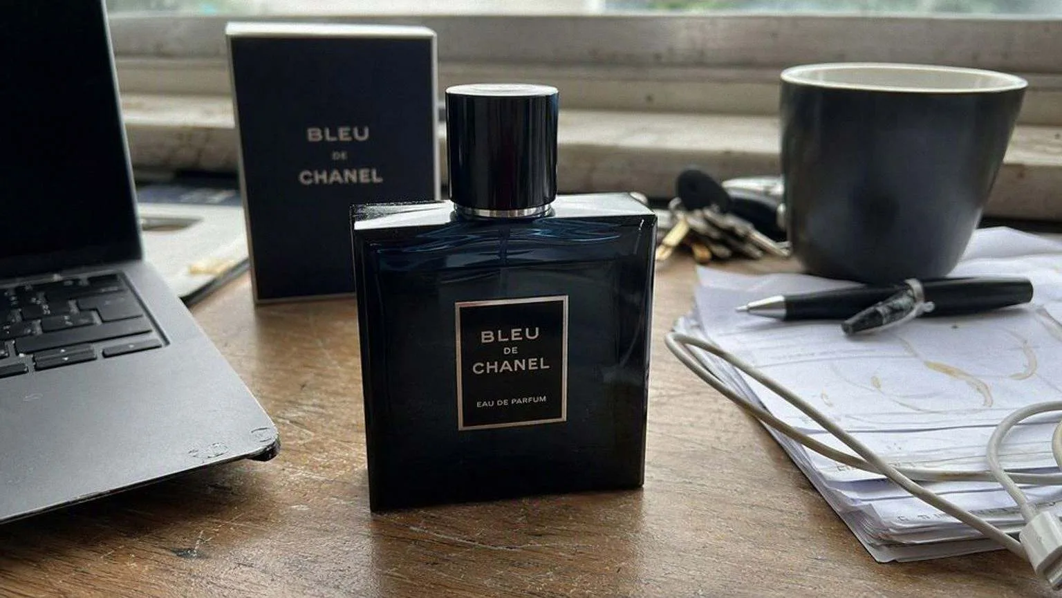 A blue fragrance bottle left on a worn wooden desk among papers, a tangled cable and a half drunk mug