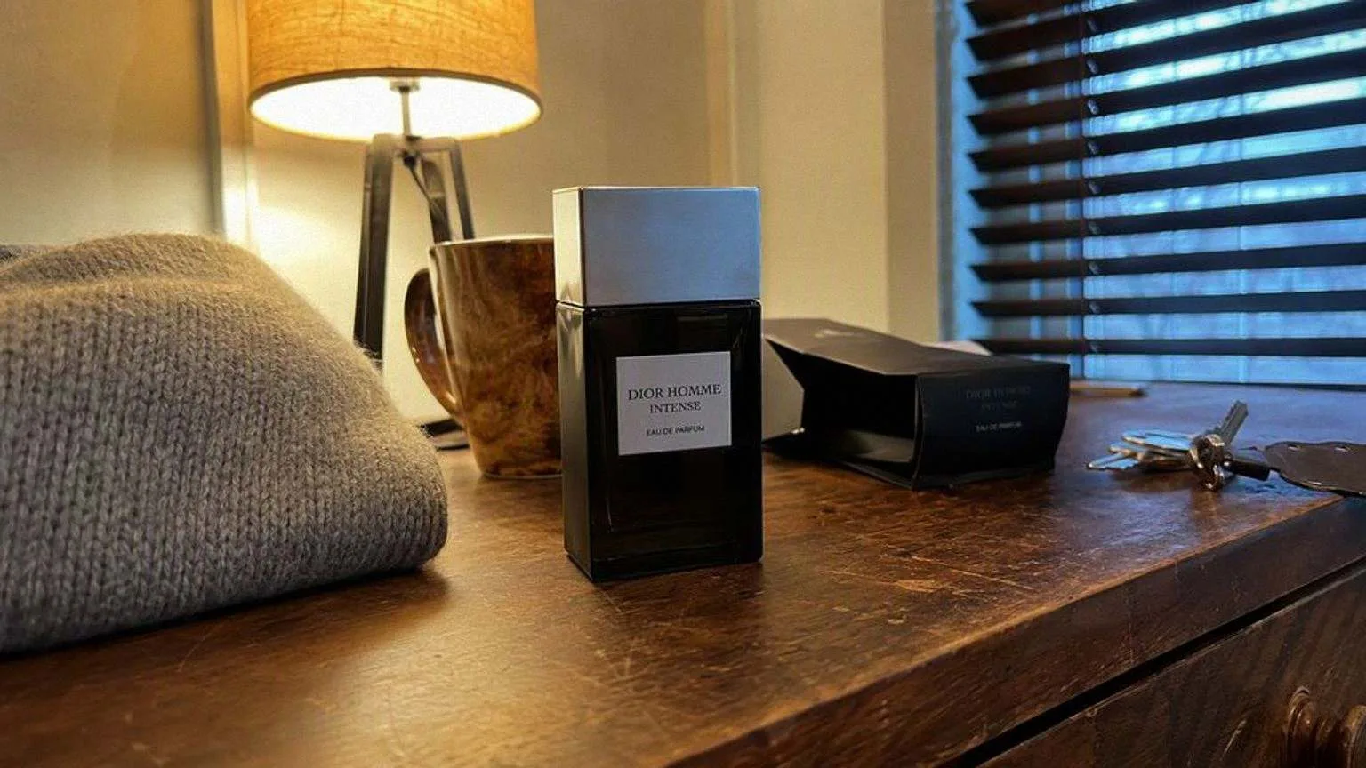 Dark Dior Homme Intense eau de parfum bottle with a silver cap on a scratched wooden dresser, a folded grey jumper beside it and a lit lamp behind