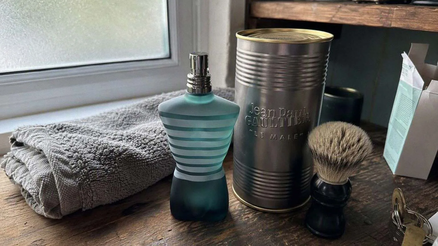 The Le Male eau de toilette bottle, a torso molded in a striped sailor shirt, standing on a tiled bathroom shelf next to a folded towel in morning light