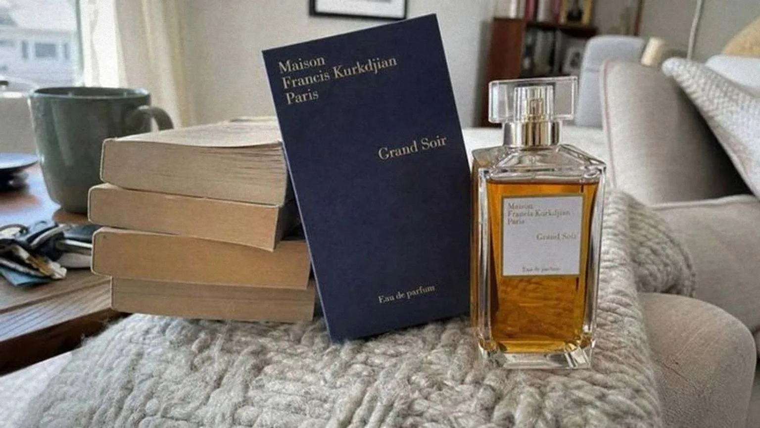The Grand Soir bottle and its navy box leaning on a stack of paperbacks, over a chunky knit blanket on a sofa arm
