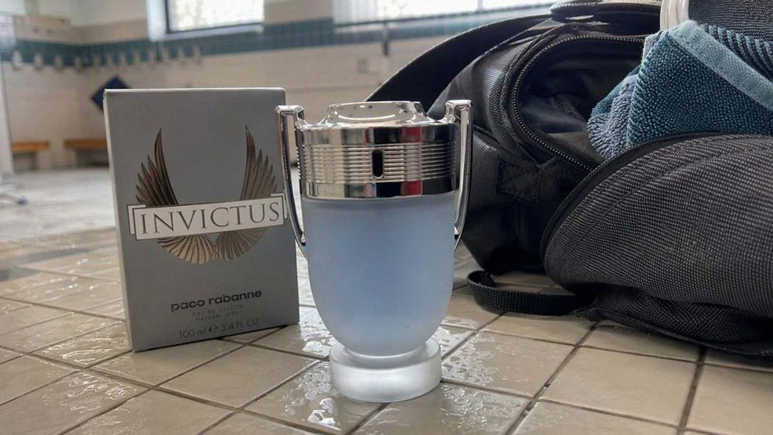 The silver and blue Invictus bottle on a wet tiled bench in a changing room, next to an open gym bag and a damp towel