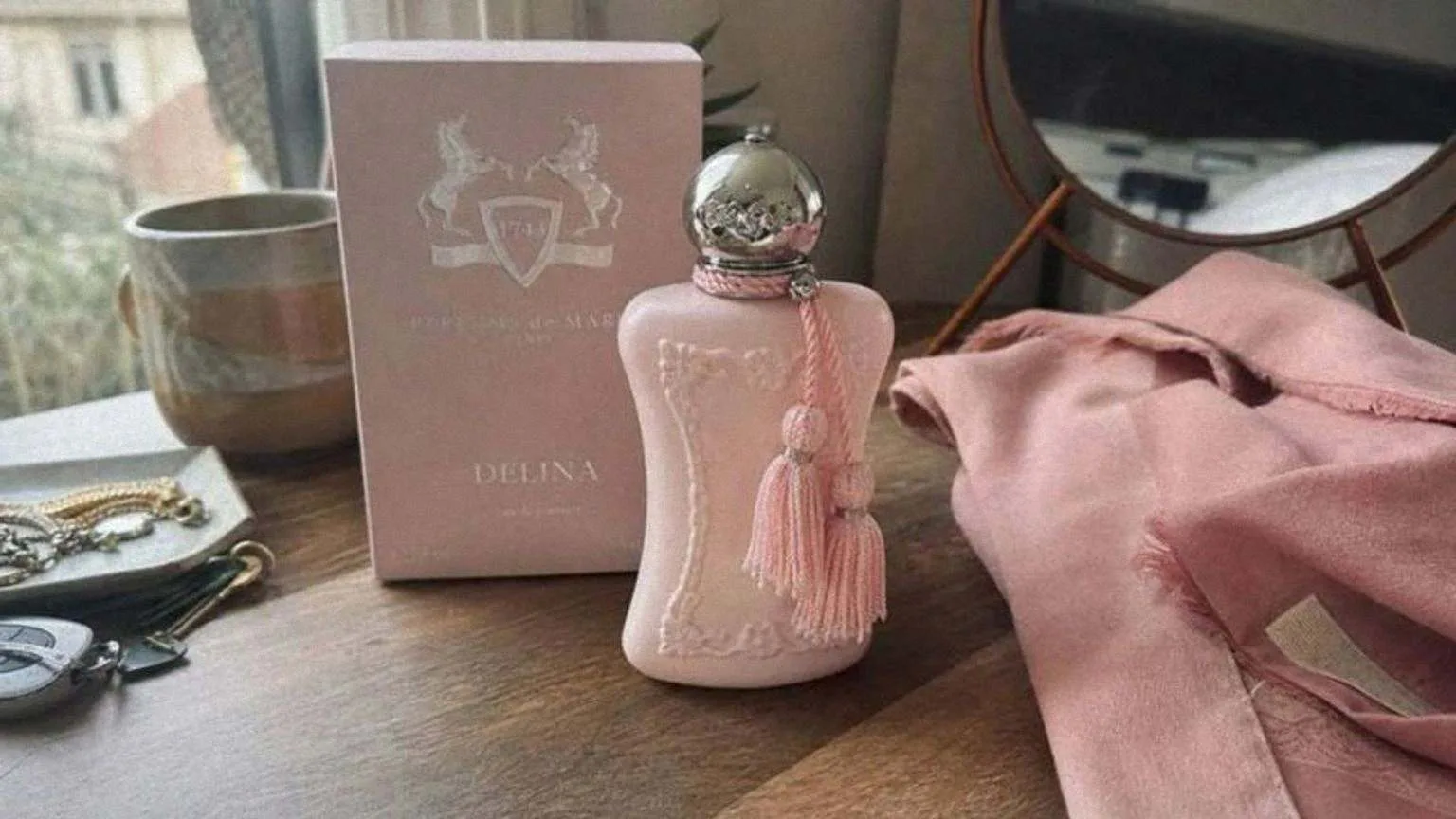 El frasco rosa de Delina con su borla, junto a la caja y un pañuelo de seda sobre un tocador de madera a la luz de la ventana