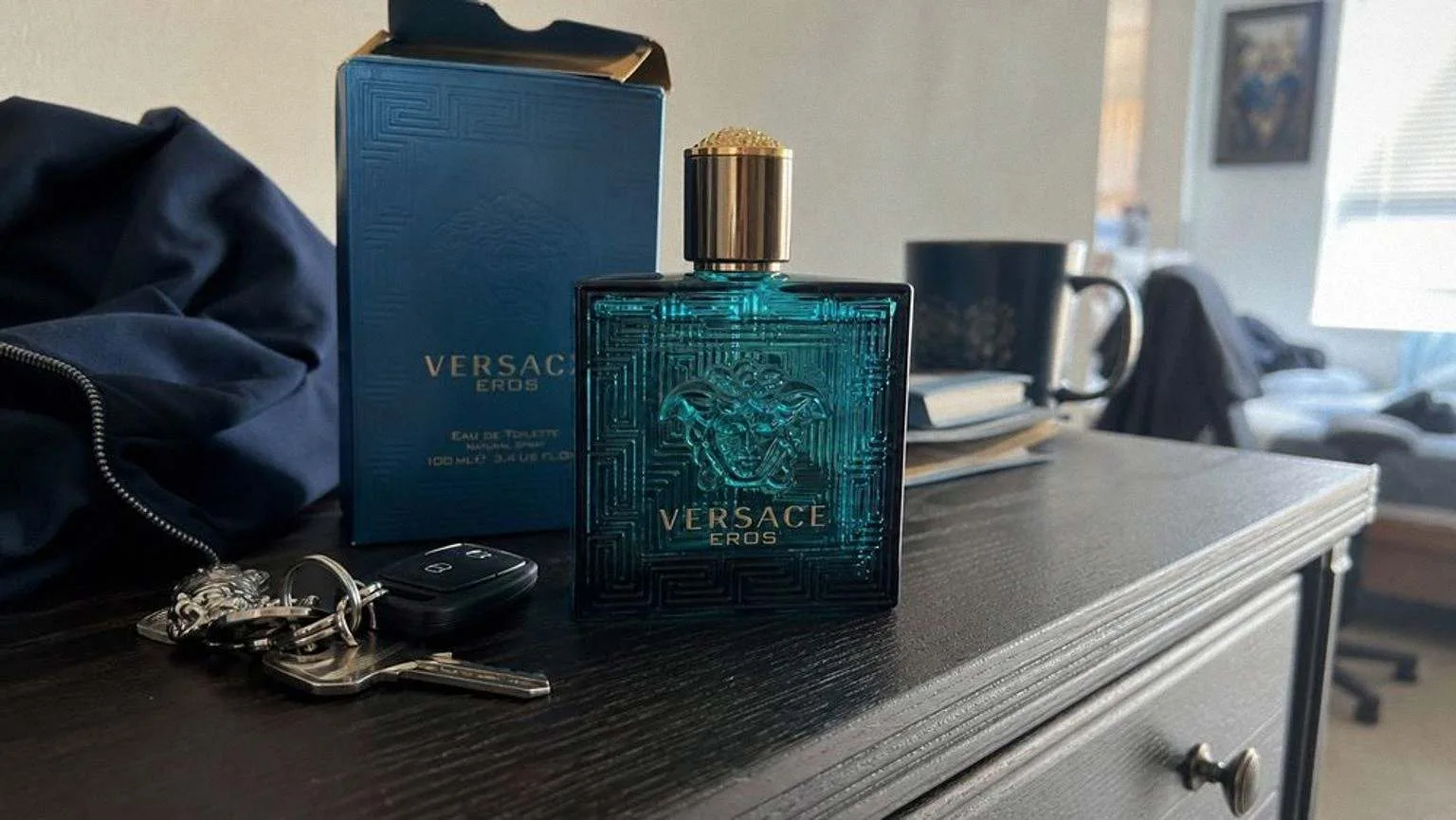 The blue Versace Eros bottle beside its box, a set of keys and a folded jacket on a dark wooden dresser